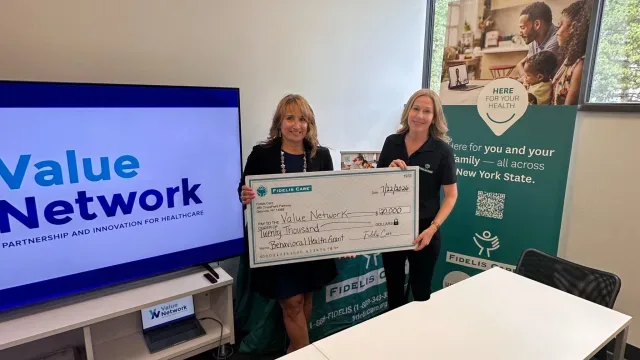 Representatives hold a Fidelis Care award check beside Value Network and Fidelis Care displays.