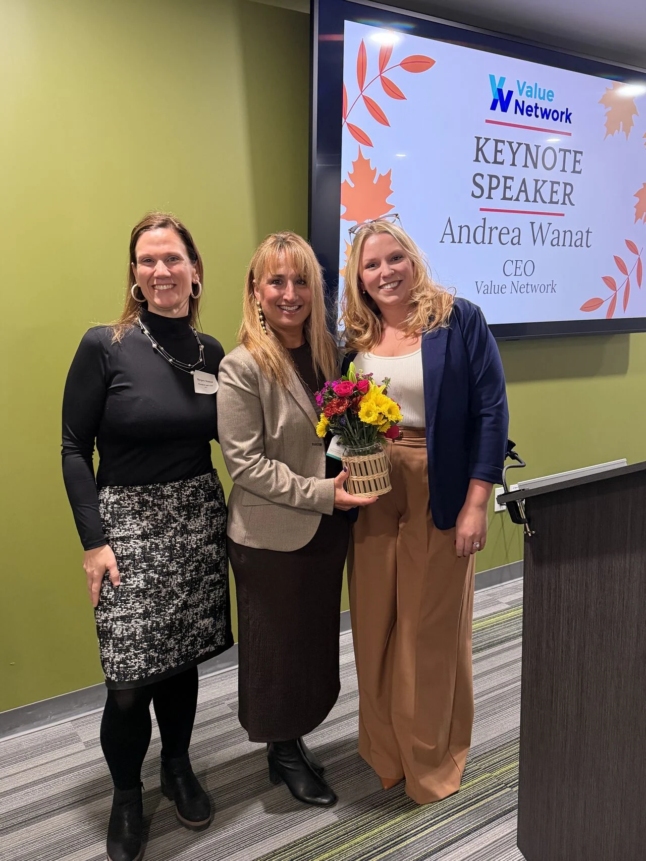 Three attendees pose beside a screen announcing keynote speaker Andrea J. Wanat.